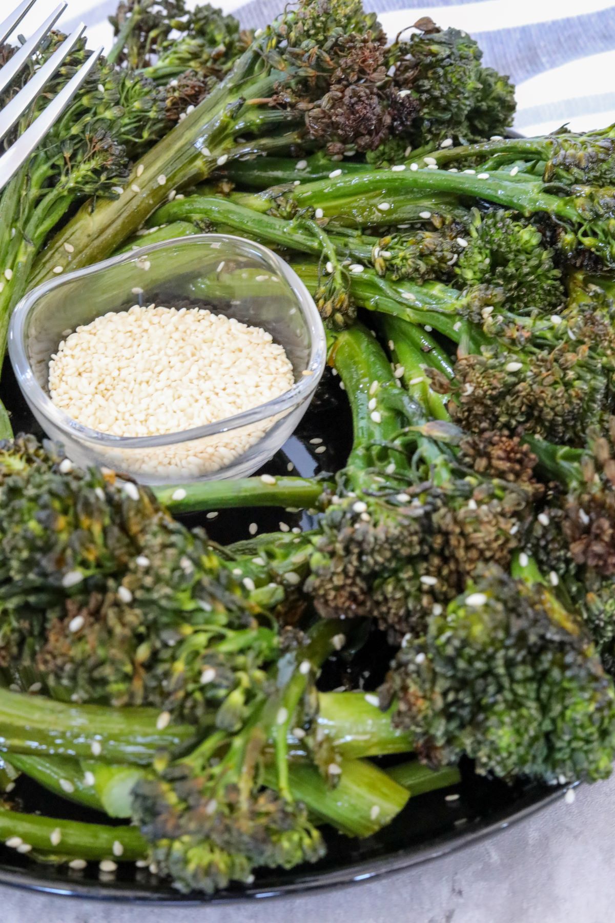close up of roasted broccolini on a black plate with a bowl of sesame seeds