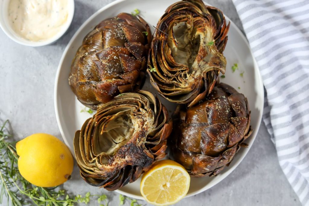 Roasted artichoke halves on a plate with a lemon half sprinkled with herbs