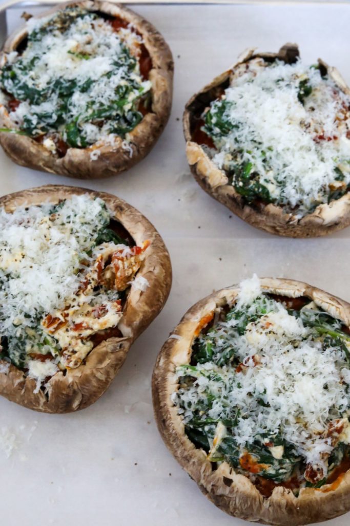 Four portobello mushrooms one a lined sheet pan topped with the spinach mixture and grated parmesan cheese