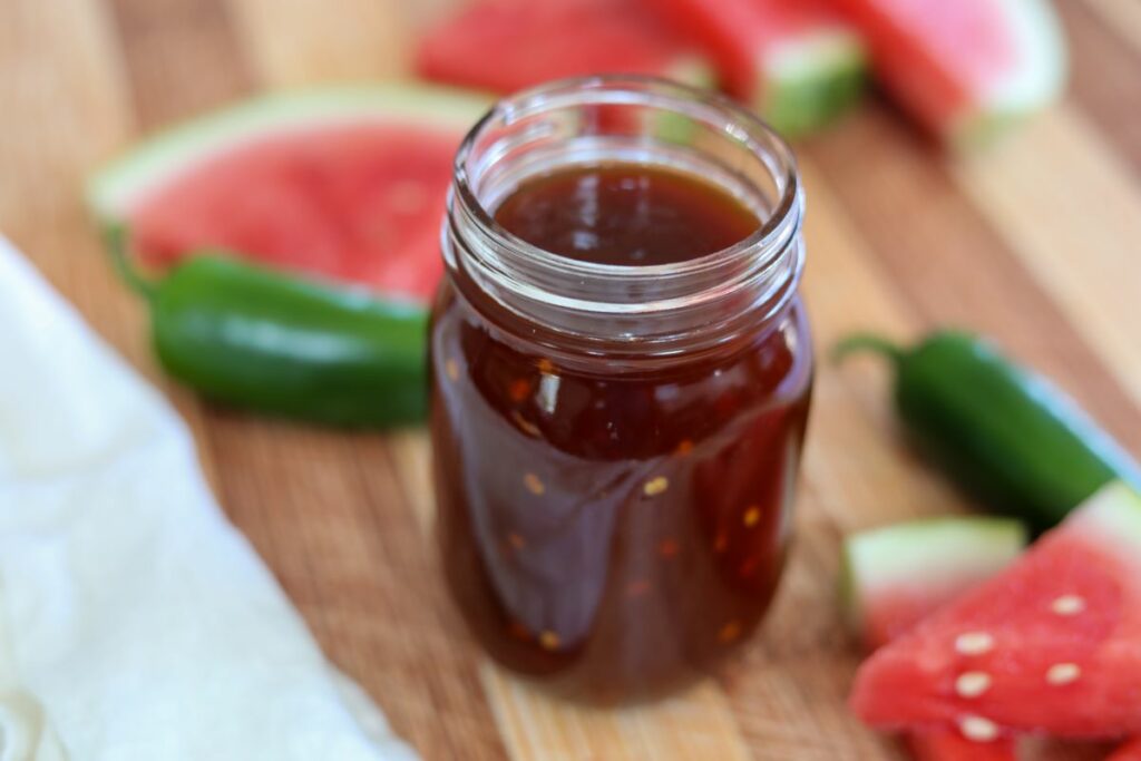 A jar of watermelon barbeque sauce surrounded by watermelon slices and jalapenos
