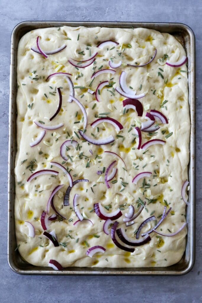 Focaccia dough in a sheet pan topped with onions, garlic and rosemary
