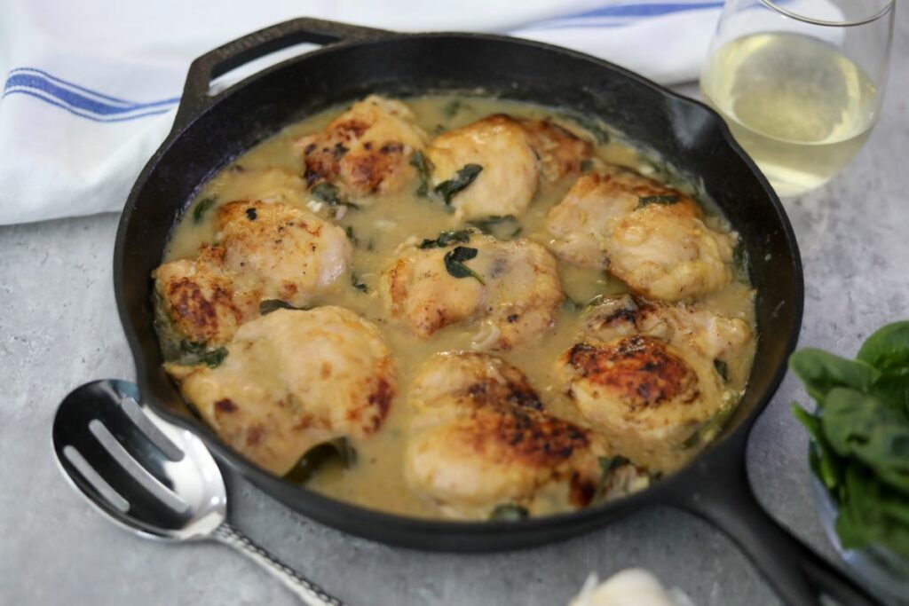 A cast iron skillet with chicken in white wine and a glass of wine
